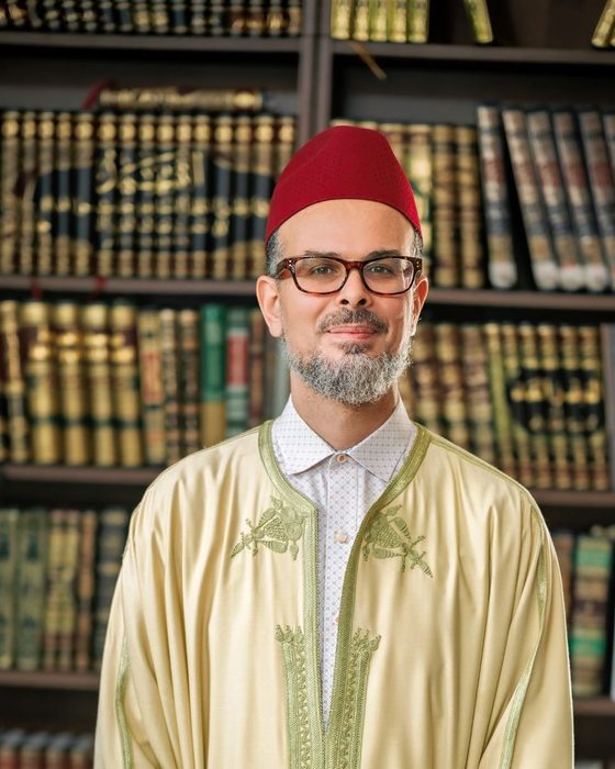 Portrait of Dr. Hassan Lachheb wearing a red fez and cream Moroccan robe, standing before shelves of classical Islamic texts